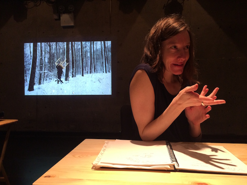 Aynsley Vandenbroucke sits in a darkened room at a desk. She looks like she is talking to someone. A video of her walking through the snowy woods with a table on her back is on in the background.